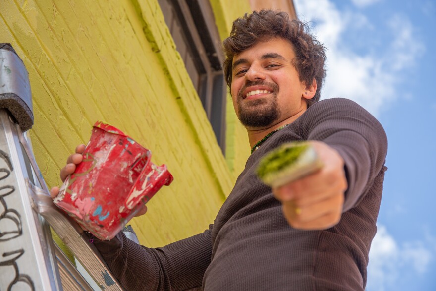 A hispanic man with short brown hair and a goatee stands up on a ladder with a smile. He's holding a paintbrush dripping with green paint out in front of him.