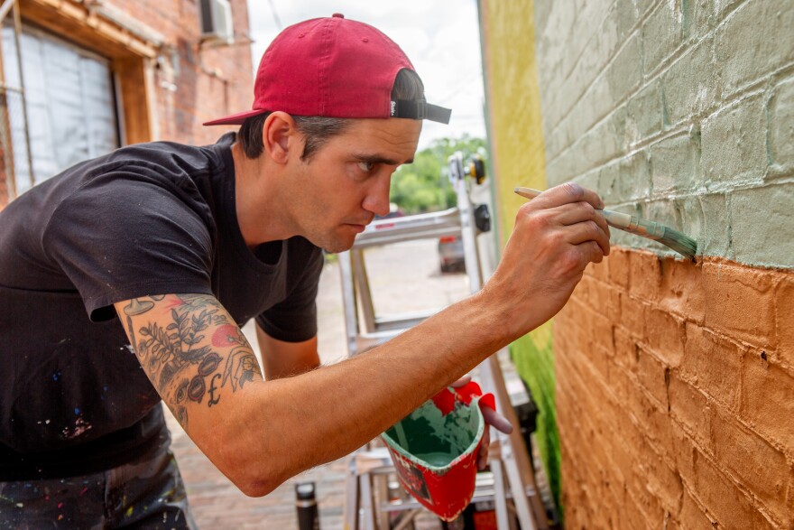 A man wearing a red backwards baseball cap and paint splattered black t-shirt leans in close to a brick wall with a thick paintbrush and brushes a greenish gray color onto the bricks.