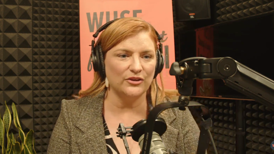 women with red hair sits and talks into a radio microphone in a studio setting