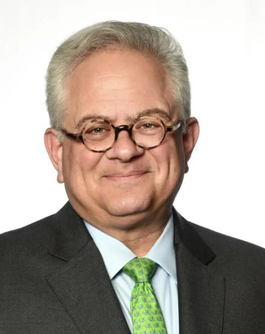 Man with combed-back gray hair, round glasses and dark suit with a yellow tie smiling into the camera