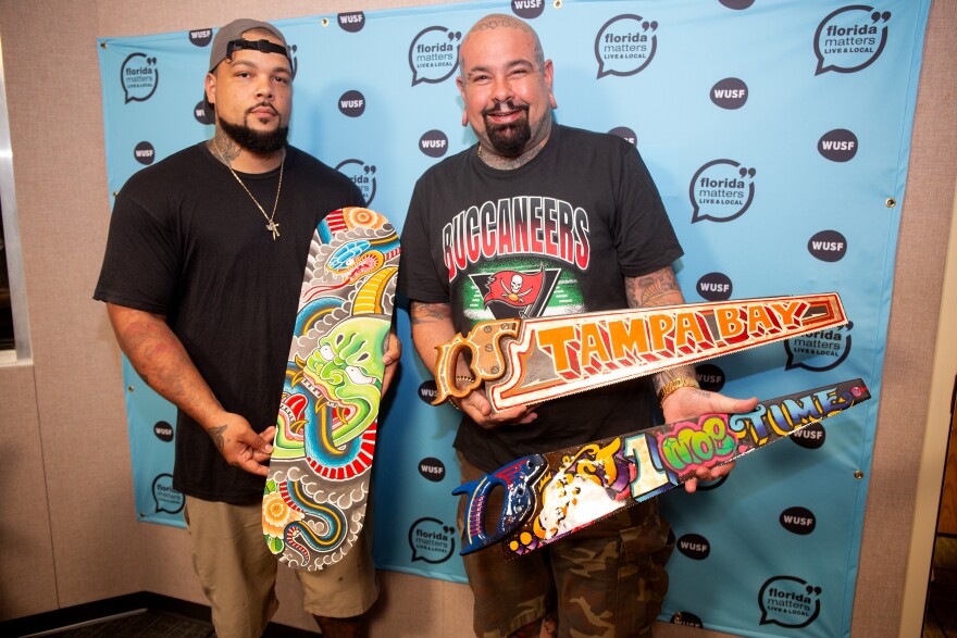 Two men wearing dark clothiung stand in front of a light blue and beige wall with the Florida Matters Live & Local logo. The man on the left is holding a skateboard deck with a colorful design on it. The  man on the right is holding two hand saws, one witch a design that says "Tampa Bay" in artistic lettering and the other which says "Just one more time" in artistic lettering. 