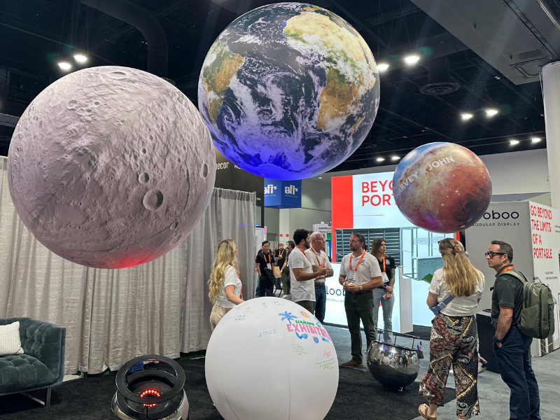 Exhibit inside exhibit hall showcases large floating planets above attendees standing and talking.