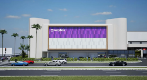Rendering of the new Wayfair store at The Galleria Fort Lauderdale featuring a modern curved building with a large Wayfair sign, glass storefront, palm trees, and cars along the adjacent roadway.