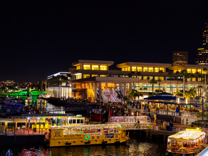 Tampa Convention Center lit up at night on a vibrant marina with lit up water taxis, a circular waterfront bar along the Tampa Riverwalk and lit up city bridges and buildings in the background.