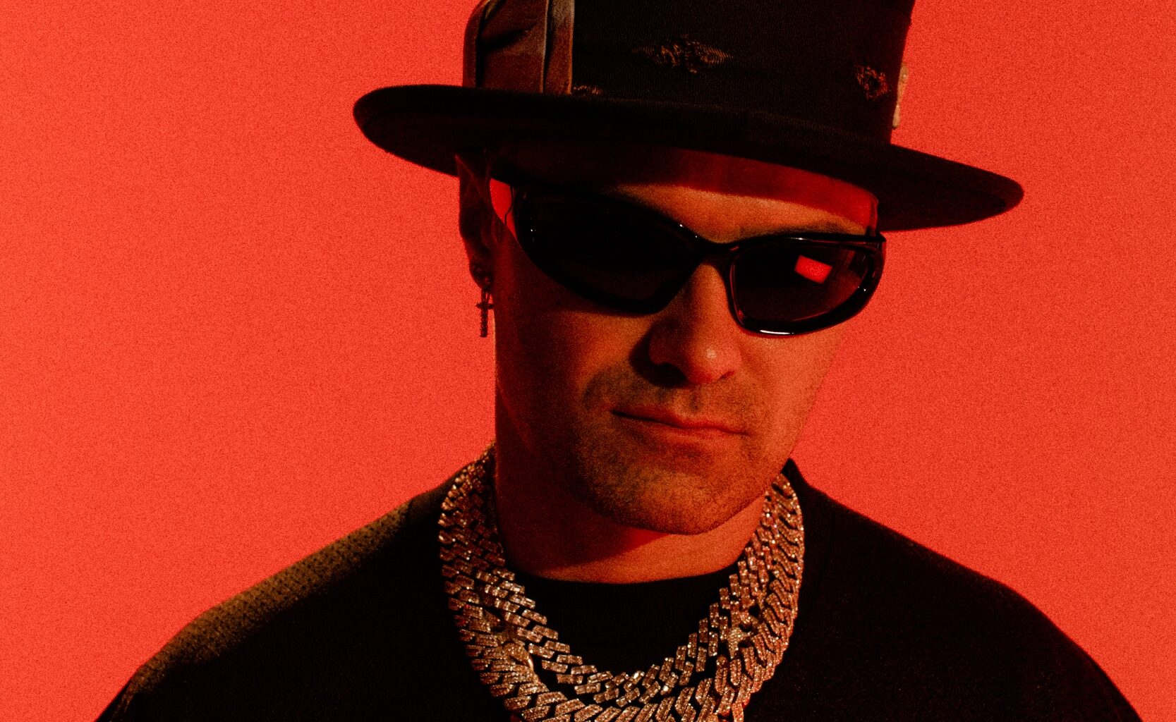photo of artist Alec Monopoly posing in a top hat, thick necklaces, and shades against a bright-red backdrop