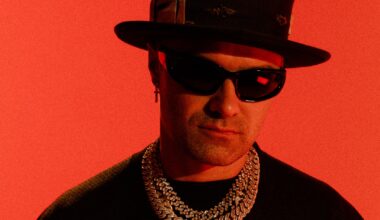 photo of artist Alec Monopoly posing in a top hat, thick necklaces, and shades against a bright-red backdrop