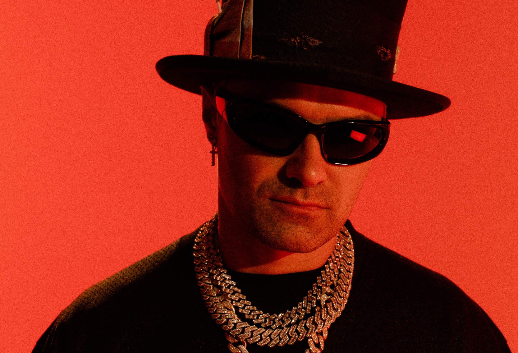 photo of artist Alec Monopoly posing in a top hat, thick necklaces, and shades against a bright-red backdrop