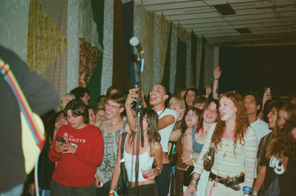 Photo of a crowd screaming and smiling as they look upon a performer on a stage. The camera shows the perspective from the stage including a microphone and the arm and guitar of a musician
