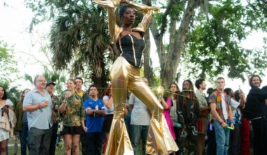 photo of a person in a black and gold bodysuit walking a makeshift runway in front of a crowd outside surrounded by trees