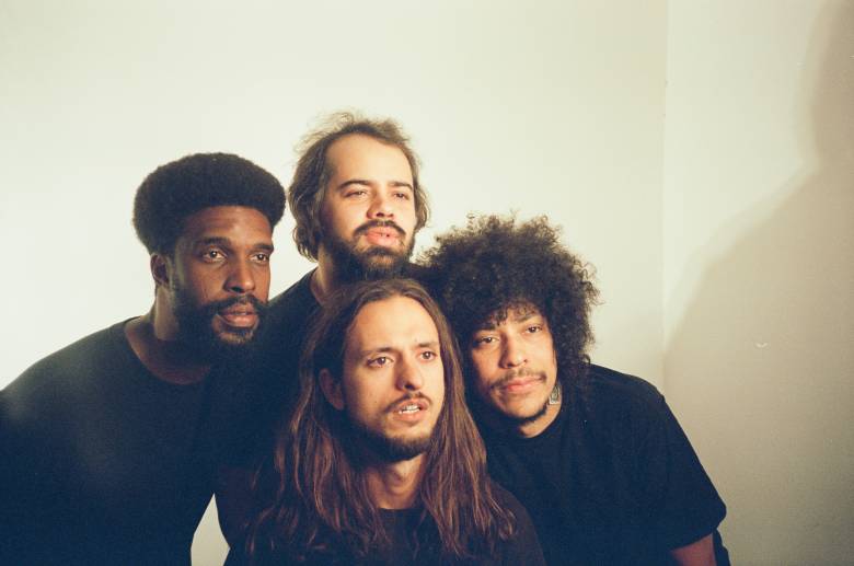A close-up group portrait of the four members of the Brazilian band Boogarins against a plain off-white wall. The band members are huddled closely together, looking directly at the camera with serious but calm expressions. From left to right: a man with a short afro and beard; a man with shorter hair and a mustache in the back; a man with long, wavy brown hair in the center foreground; and a man with a large, voluminous afro on the right. They are all wearing dark clothing, and the lighting is warm and soft, casting gentle shadows against the corner of the wall.