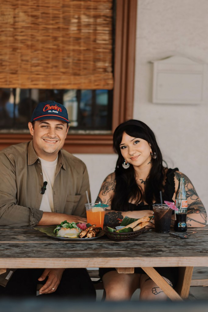 Nate Siegel, founder of Cheeky’s, and Chef Julie Sainte Michelle Feliciano, founder of Lucky Tigre, sitting at a wooden outdoor table during a pop-up event. Nate wears a "Cheeky’s St. Pete" hat, while Julie is seated next to a spread of Filipino cuisine, including lumpia and colorful tropical cocktails.