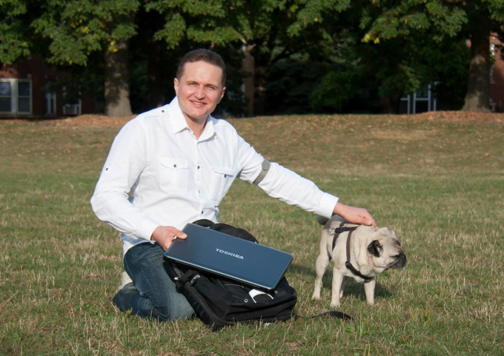 A photo of a man in a white button down and jeans in the grass with one hand holding a laptop and another petting a Pug. 