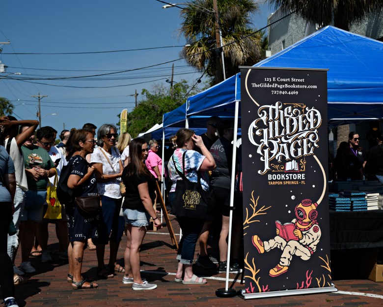 People standing in line at Tarpon Springs Book Festival outdoor event in front of a tent with a sign for The Gilded Page bookstore.