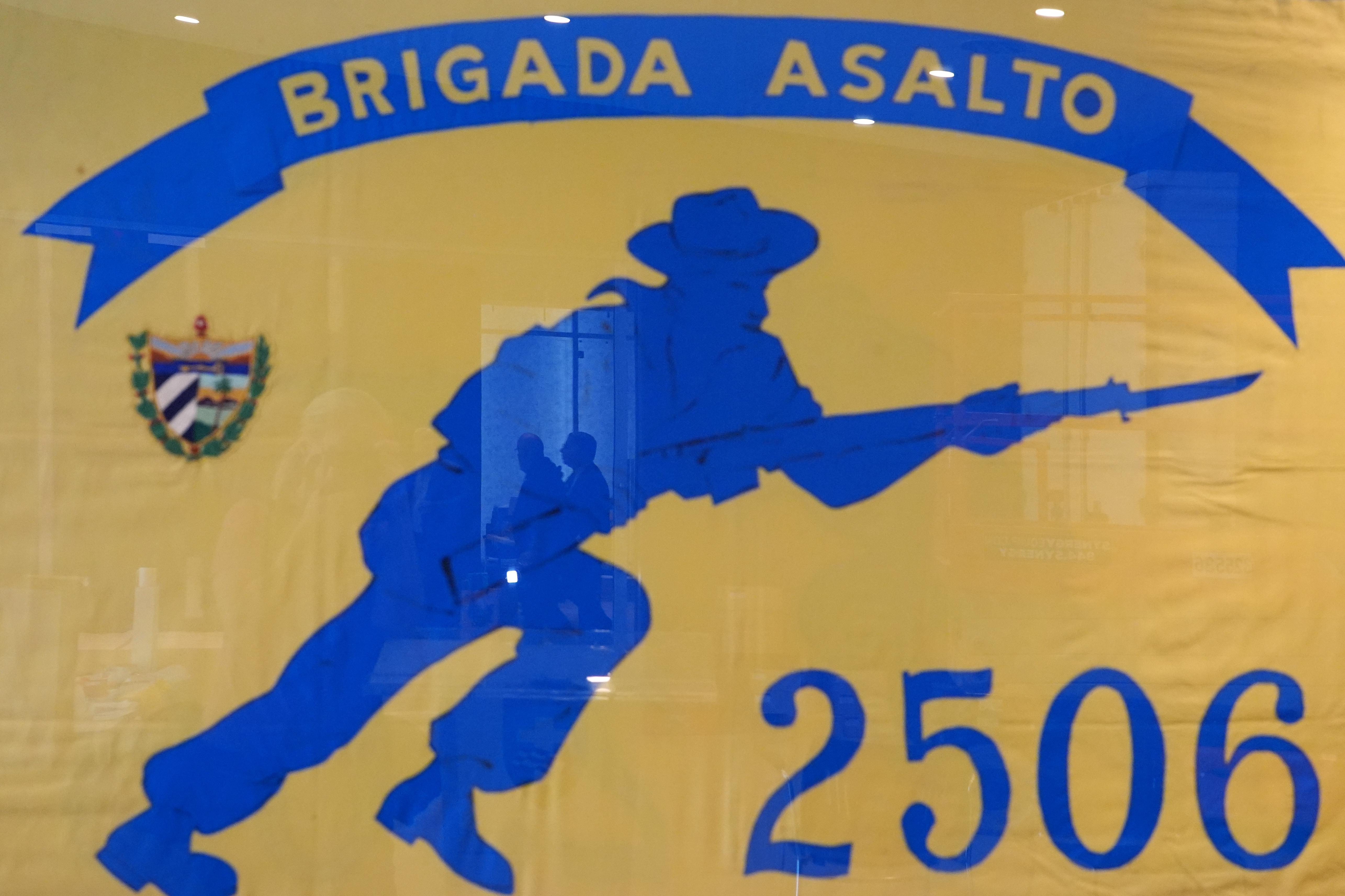 People are reflected in a framed Brigade 2506 flag, as...