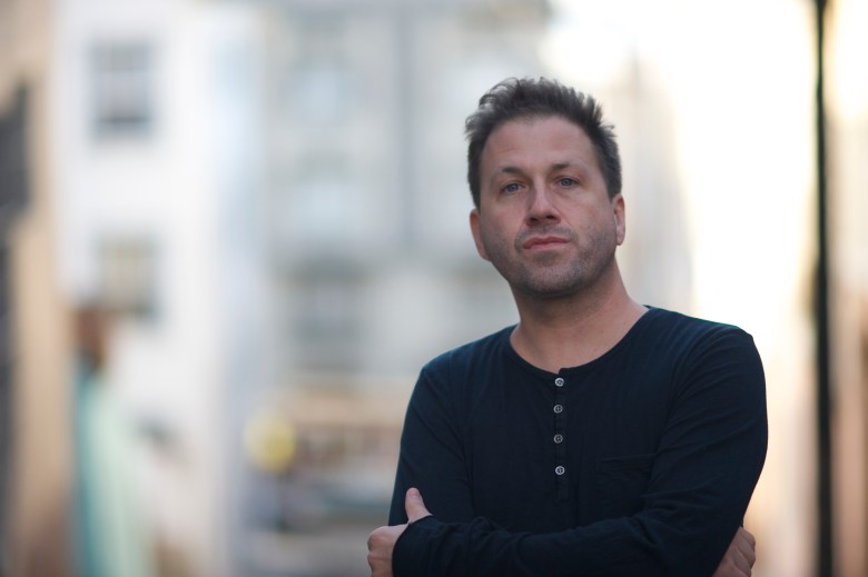 A professional portrait of DJ Three (Christopher Milo) posing outdoors. He is wearing a black long-sleeved henley shirt and has his arms crossed, looking directly at the camera with a calm expression. The background is a soft-focus urban street with light-colored buildings under bright, natural light.