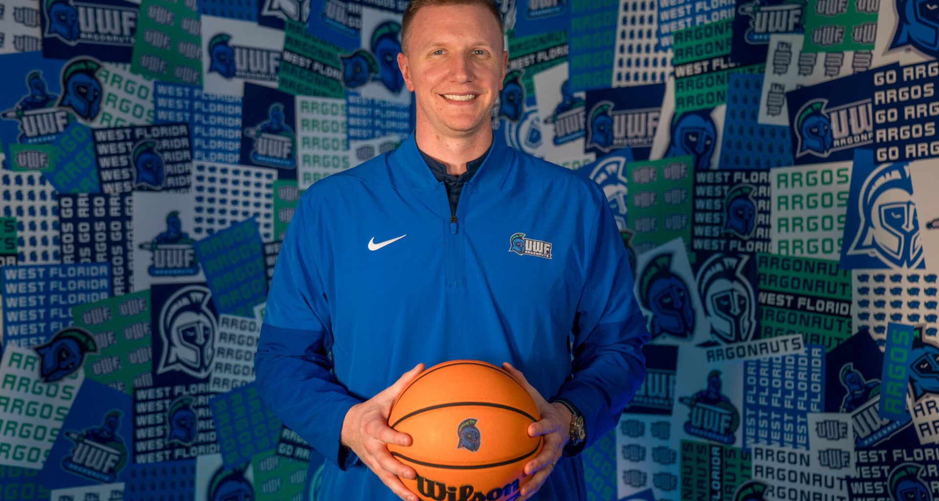 Basketball coach holds basketball in hands in front of a collage wall with Argo graphics.