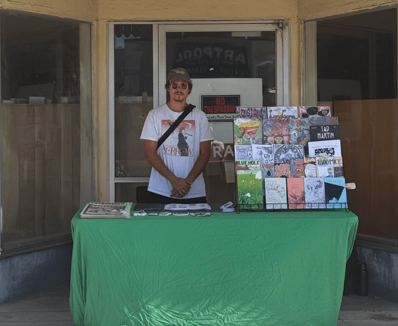 Filipe Bergson stands behind a green-clothed table set up in the recessed entryway of Haslam’s Book Store. He is wearing a white graphic t-shirt, a baseball cap, and a crossbody bag. The table is lined with a wire rack displaying various indie comic books and zines. The darkened windows and closed doors of the historic bookstore serve as the backdrop under natural outdoor lighting.