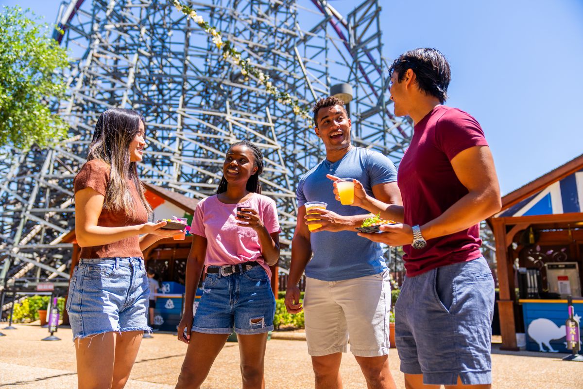 Busch Gardens Tampa Bay theme park guests are seen talking in front of a large roller coaster, beverages in hand.