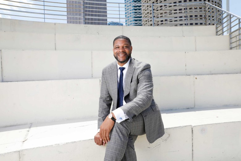 A professional portrait of Gary Hartfield sitting on white concrete bleacher-style steps in an urban park. He is smiling and wearing a grey plaid suit with a navy blue tie and a white dress shirt. The Tampa city skyline and a metal railing are visible in the blurred background under bright daylight.