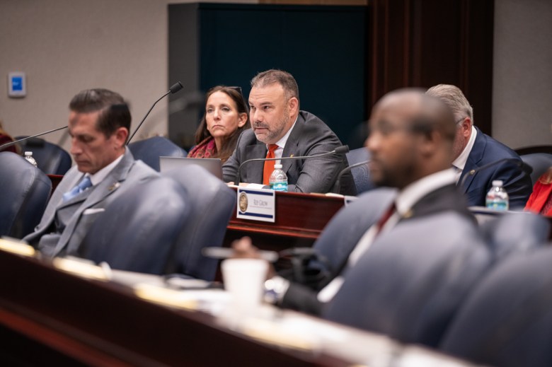 In a formal hearing room, several people are seated in rows. The central focus is on a man with a beard, wearing a grey suit and red tie, looking forward attentively.