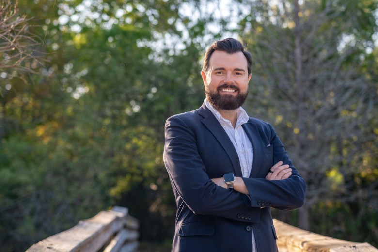 A professional outdoor portrait of Hoyt Prindle III. He is smiling and standing with his arms crossed, wearing a navy blue blazer over a light-colored button-down shirt. He has dark hair and a groomed beard. The background is a soft-focus view of a wooden bridge and green trees under natural daylight.