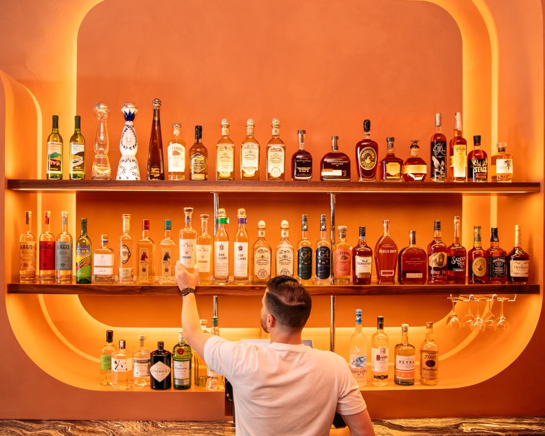 A bartender reaching for a bottle of liquor from a brightly lit, retro-style bar shelf glowing with warm orange light at Easy Tiger in St. Petersburg.