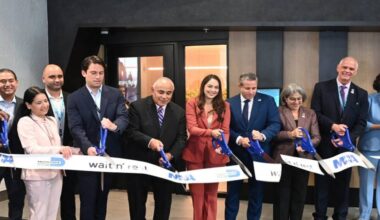 A group of officials and business leaders stand in a row indoors, smiling as they cut a ceremonial ribbon at Miami International Airport with oversized blue scissors.