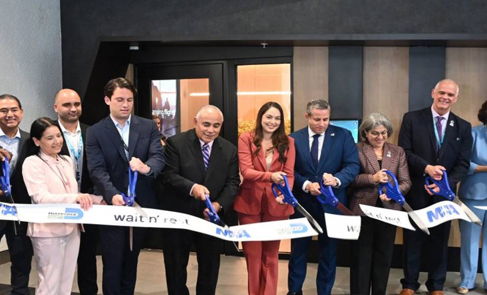 A group of officials and business leaders stand in a row indoors, smiling as they cut a ceremonial ribbon at Miami International Airport with oversized blue scissors.