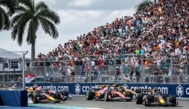 Formula 1 cars race around a track corner as a large crowd of spectators watch from the grandstands under palm trees on a sunny day. Advertising banners line the fence alongside the track.
