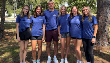 Six students stand together in matching blue t-shirts, smiling, with trees behind them.