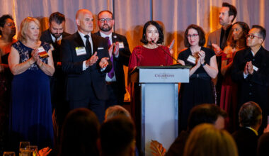 A wide shot of a formal awards ceremony or gala. A woman with dark hair, wearing a red long-sleeved dress, stands at a light blue podium labeled "CHAMPIONS" and speaks into a microphone. To her left and right, several colleagues in suits and formal dresses stand on stage, clapping and looking toward her with supportive expressions. The background features warm, draped gold curtains, and the blurred silhouettes of an audience are visible in the foreground.