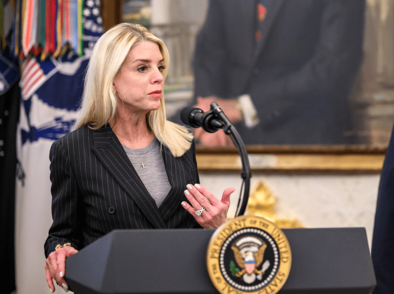 A medium shot of Attorney General Pam Bondi speaking at a dark grey presidential podium inside the Oval Office. Bondi has long blonde hair and is wearing a dark, pinstriped blazer over a grey top. She is gesturing with her right hand while speaking into a dual-microphone setup. The podium features the gold-and-blue circular Seal of the President of the United States.