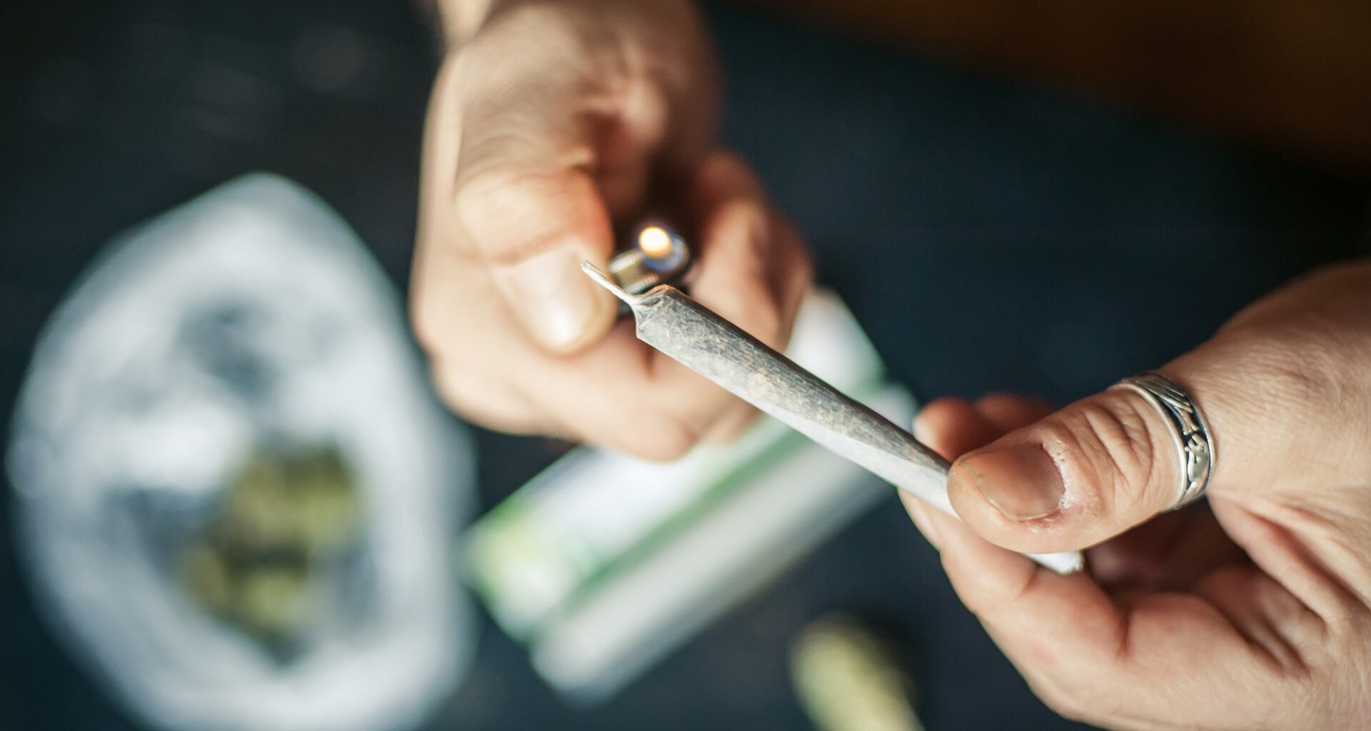 photo of a closeup of hands lighting a marijuana joint with marijuana on the table in a baggie on a table below