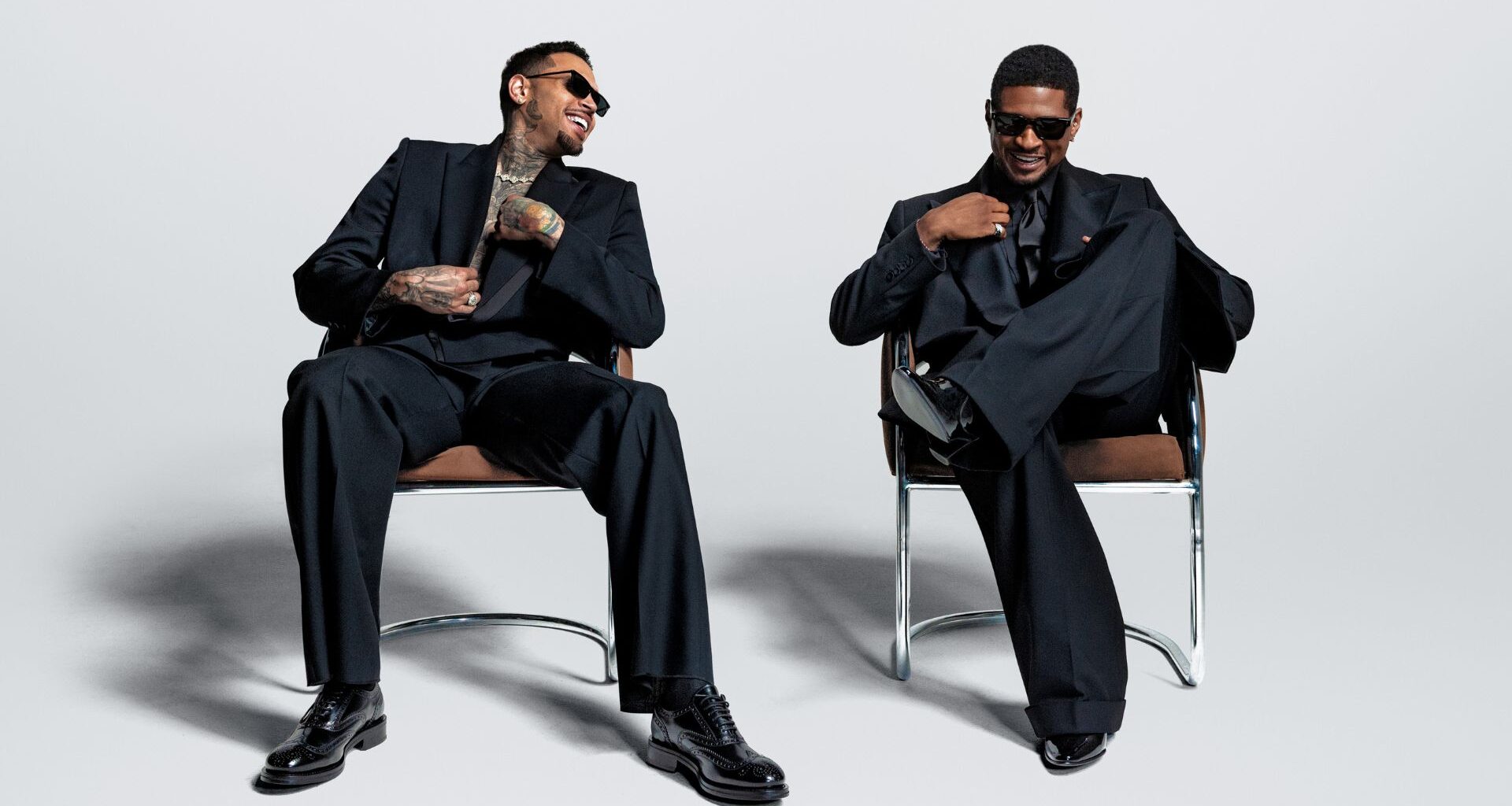 Photo of Usher and Chris Brown sitting on chairs wearing black suits.