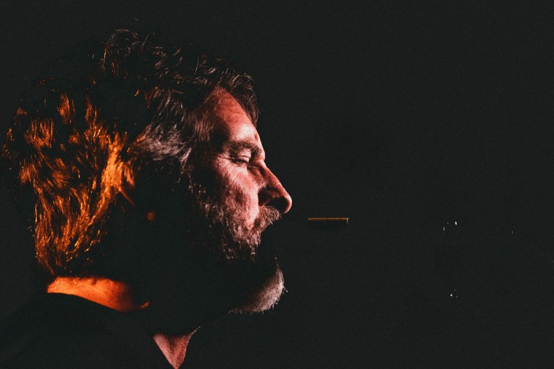 A dramatic side-profile portrait of Addie Burns from the band QUIT. He is captured in a dark, low-light setting with warm orange light highlighting his hair, beard, and the side of his face against a black background. He appears to be singing or speaking into the darkness.