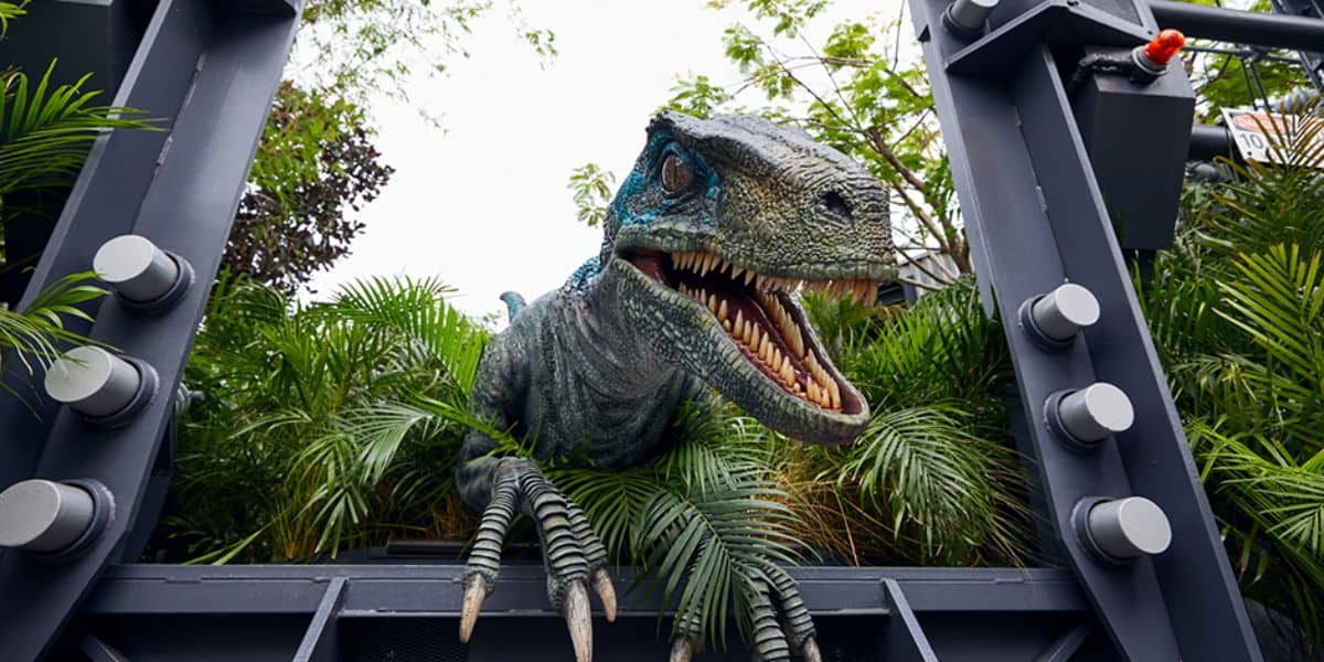 A large, menacing velociraptor with an open mouth and sharp teeth emerges from dense greenery and mechanical structures at the Jurassic Park Tribute Store in Universal, set against a background of trees and foliage. The dinosaur appears life-like and intimidating.