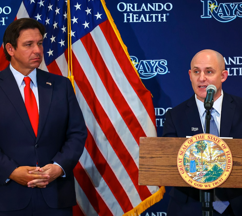 Governor Ron DeSantis (left) stands in a dark suit and red tie, looking on as Ken Babby (right) speaks from a wooden podium bearing the Great Seal of the State of Florida. The background consists of a blue step-and-repeat backdrop with "Orlando Health" and "Rays" logos, flanked by a large American flag.