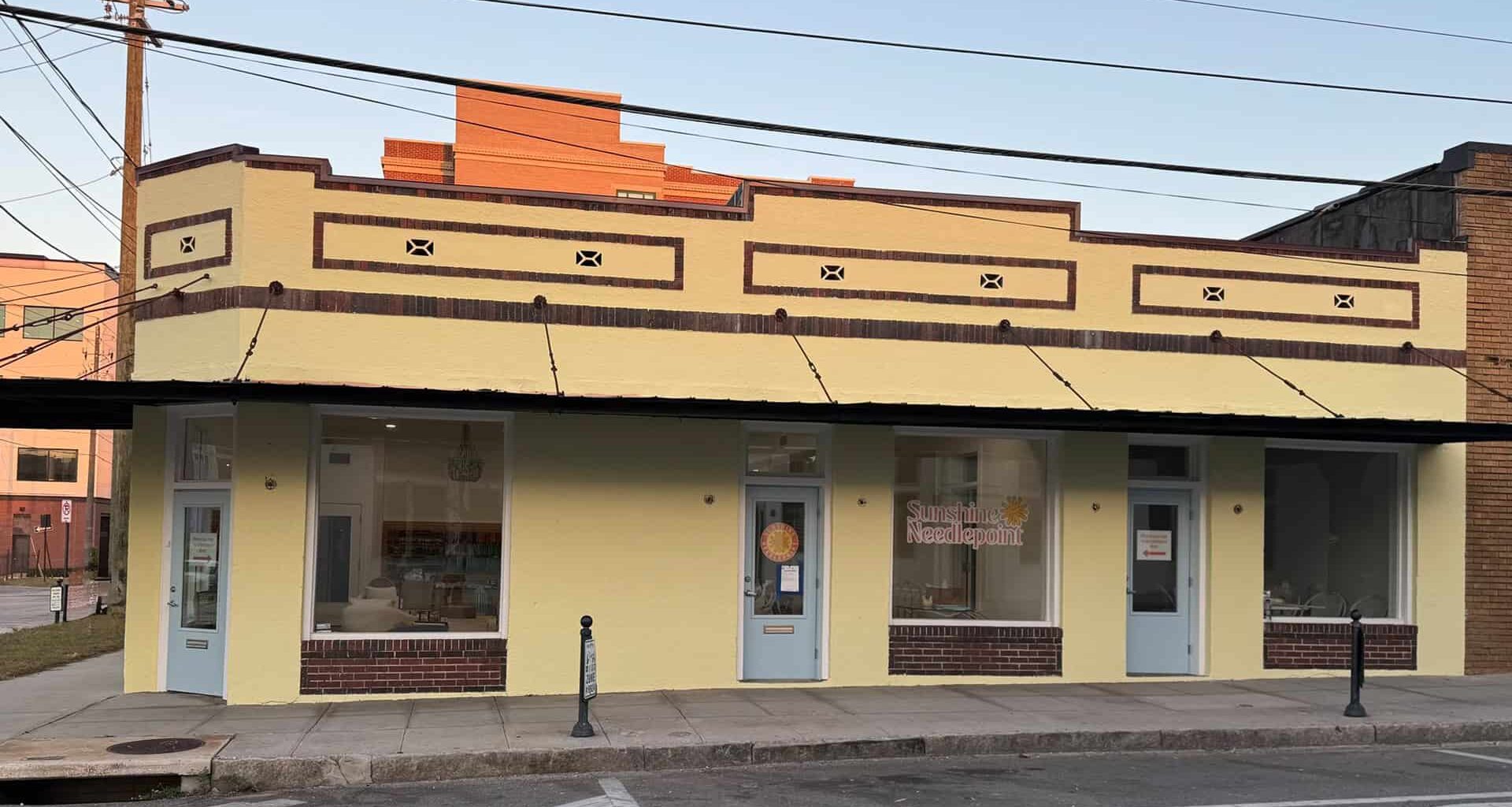 Photograph of a single-story yellow building with brown trim, featuring three large windows, three doors along a sidewalk, and signage for “Sunshine Needlepoint”.