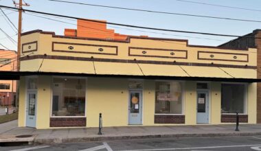 Photograph of a single-story yellow building with brown trim, featuring three large windows, three doors along a sidewalk, and signage for “Sunshine Needlepoint”.