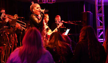 A female singer performs on stage at Seminole Classic Casino under red and purple lighting, holding a microphone and singing energetically while a live band with brass instruments plays behind her. Audience members are visible in the foreground, watching the performance.