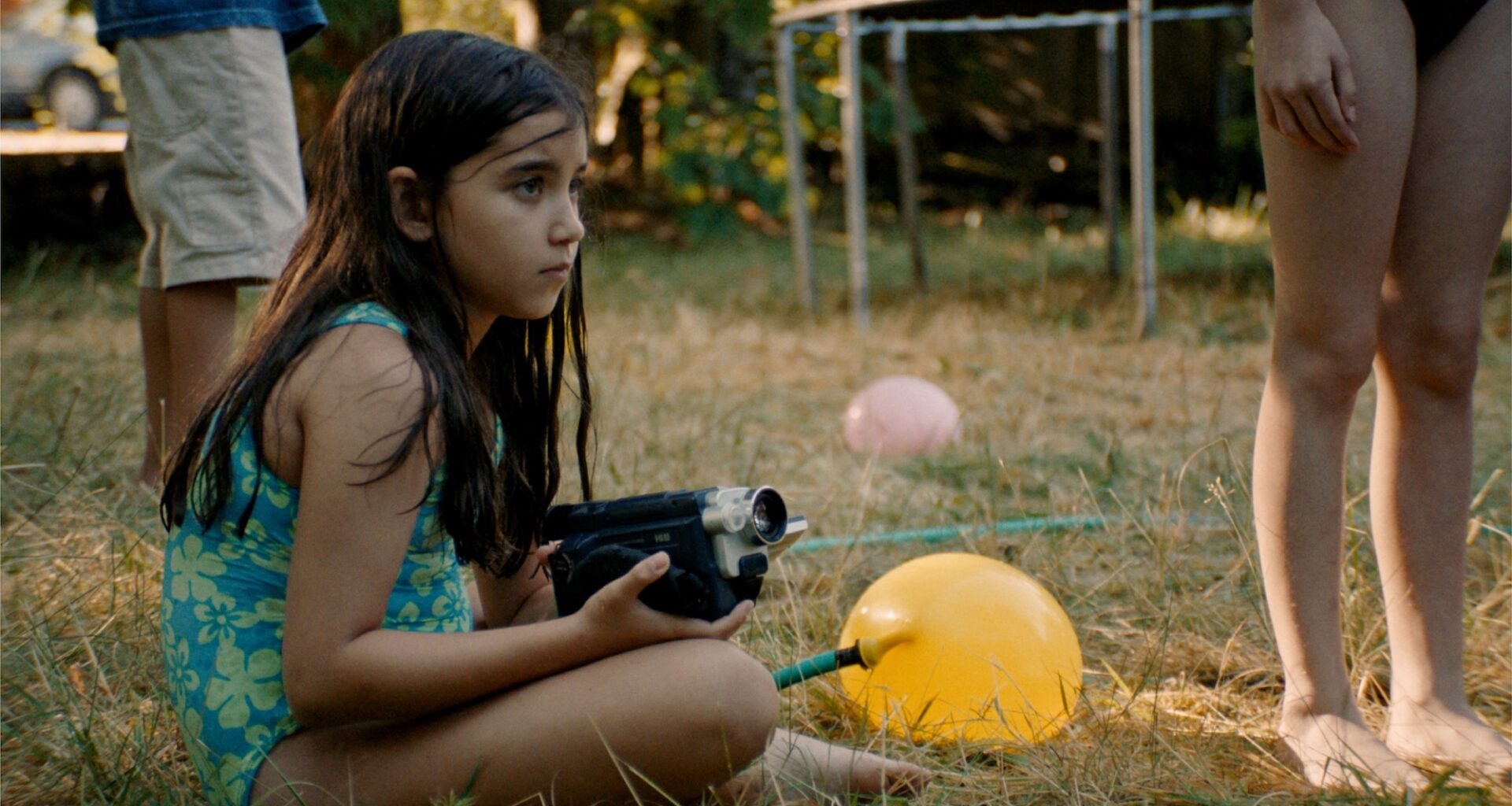 still from the movie 'Blue Heron' showing a young girl in a bathing suit sitting on the grass and holding a camcorder