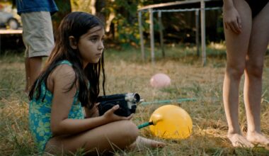 still from the movie 'Blue Heron' showing a young girl in a bathing suit sitting on the grass and holding a camcorder