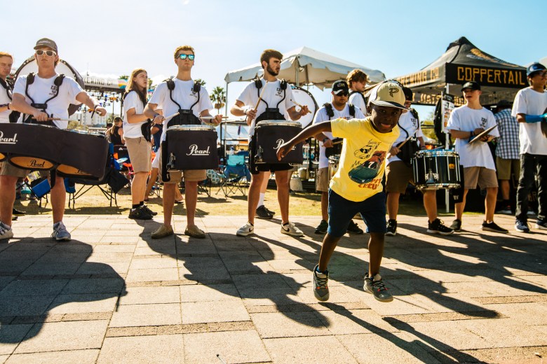 A young boy in a yellow t-shirt and a baseball cap dances joyfully on a stone-paved walkway at the Gasparilla Music Festival. Behind him, a high school drumline in white t-shirts and blue shorts performs with marching snares and tenors. A "Coppertail Brewing Co." tent and festival goers are visible in the sunny background.