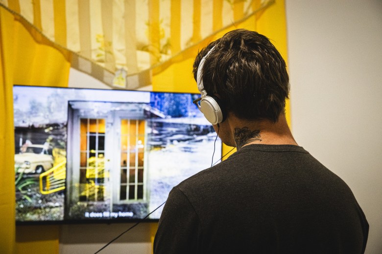 A gallery visitor seen from behind, wearing over-ear headphones and watching a video installation on a flat-screen monitor. The screen displays a grainy image of a small building at night. A yellow and white striped fabric draped with a gold chain hangs behind the monitor on the white gallery wall.