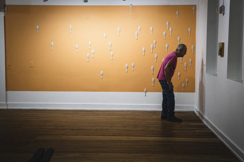 A man in a pink shirt and dark pants leans in closely to examine a small, framed artwork on a white gallery wall. To his left, a large, rectangular tan-colored installation is mounted on the wall, dotted with dozens of small, white sculptural elements. The scene is set on dark wood floors under soft gallery lighting.