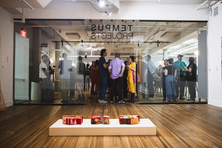 A wide interior shot of the Tempus Projects art gallery in Ybor City during an opening reception. In the foreground, three small colorful sculptures sit on a white pedestal. In the background, a crowd of people is visible through a glass partition labeled "TEMPUS PROJECTS" in reverse, with guests mingling in a brightly lit, modern industrial space.