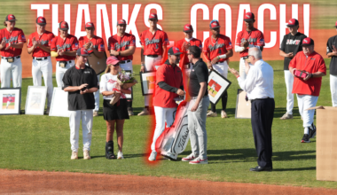 Baseball team on a field presenting awards; a coach receives flowers and a framed photo as players applaud, with a large 'THANKS COACH' banner above.