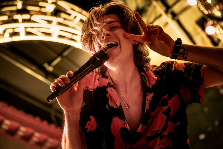 A dynamic, close-up shot of William Beckett, lead singer of The Academy Is..., performing live on stage. He is captured in a moment of high energy, leaning into a microphone held in his right hand while flashing a peace sign with his left. He has mid-length, wavy brown hair falling over his face and is wearing a dark button-down shirt featuring a bold, pink floral pattern. The lighting is warm and golden, highlighting a tattoo on his left wrist and a delicate silver necklace. The background features the blurred architectural details of a concert venue ceiling and stage lights.
