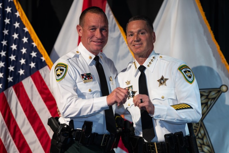 Sheriff Chad Chronister and Major Troy Morgan (left) during an official promotion ceremony. Both are dressed in white long-sleeved dress shirts with black ties and silver badges. They are standing in front of several flags, including the American flag and the Hillsborough County Sheriff's Office flag, with gold-fringed edges.