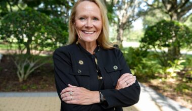 Blonde woman wearing black collared shirt poses with arms crossed outside with green trees behind her